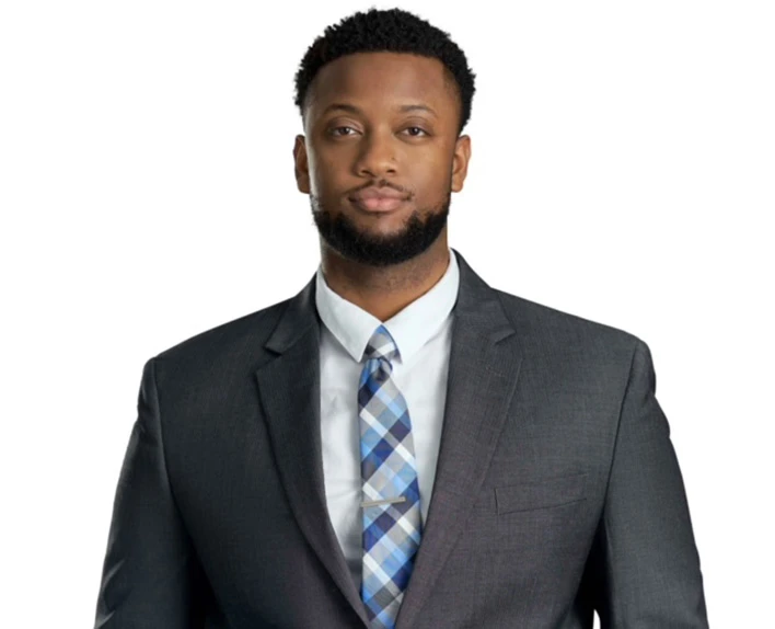 Professional man in dark suit and plaid tie standing against white background.