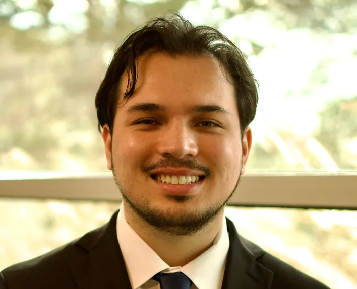 Professional man in dark suit and plaid tie standing against white background.